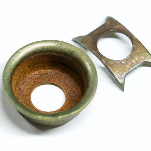 May include: Close-up of two aged metal components. One is a round, cup-shaped piece with a central hole and a green-tinged rim. The other is a flat, angular piece with a circular opening and pointed edges. Both show signs of rust.