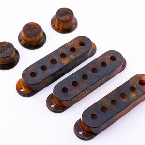 Könnte beinhalten: Satz mit vier Gitarren-Tonabnehmerabdeckungen und drei Knöpfen. Die Abdeckungen sind in einem gefleckten Braun- und Schwarzton gehalten. Die Knöpfe sind braun mit schwarzer Beschriftung. Die Knöpfe sind mit "Volume" und "Tone" beschriftet.