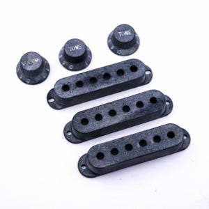 Könnte beinhalten: Eine Sammlung von Gitarrenteilen, darunter drei schwarze Tonabnehmerabdeckungen mit jeweils sechs Löchern und drei Reglerknöpfe. Die Knöpfe sind mit "VOLUME" und "TONE" mit Zahlen markiert. Die Teile sind auf einer weißen Oberfläche angeordnet.