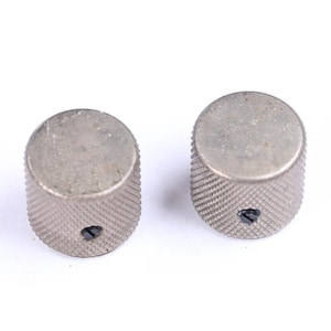 May include: Two silver-colored, knurled knobs. Each knob has a cylindrical shape with a flat top and a small hole. The knobs are likely for a musical instrument or electronic device.