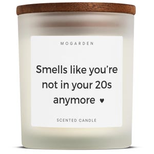 May include: A white scented candle with a wooden lid. The candle has a label that reads "Smells like you're not in your 20s anymore." The brand name "MOGARDEN" is at the top of the label.