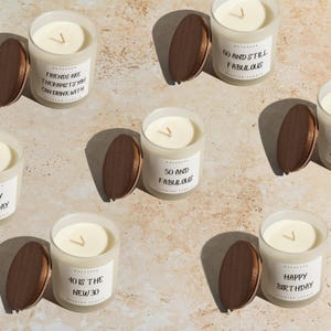 May include: Several glass candles with wooden lids are arranged on a light-colored surface. The candles have white wax and labels with phrases like "Happy Birthday" and "50 and Fabulous." The overall theme is celebratory and gift-oriented.