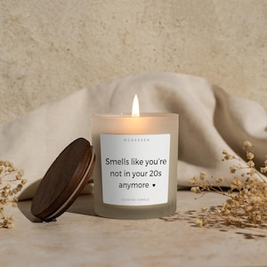 May include: A lit, frosted glass scented candle with a wooden lid. The candle has a white label with the text "Smells like you're not in your 20s anymore." The candle is next to a wooden lid and dried flowers.