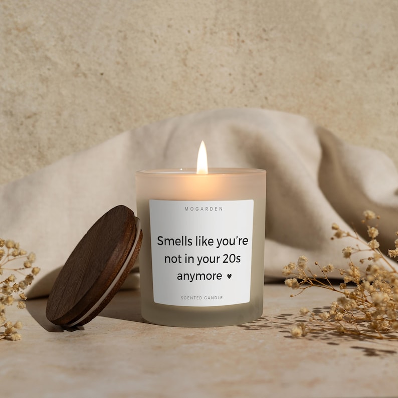 May include: A lit, frosted glass scented candle with a wooden lid. The candle has a white label with the text "Smells like you're not in your 20s anymore." The candle is next to a wooden lid and dried flowers.