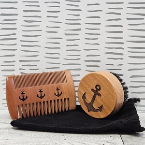 May include: A wooden beard comb and brush set with an anchor design. The comb has three anchors carved into the wood and the brush has one anchor carved into the wood. The set comes with a black velvet pouch.