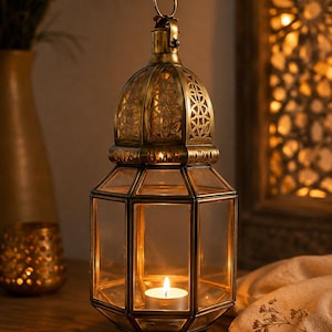 May include: A hexagonal lantern crafted from antique brass-toned metal and clear glass panels. The top features an intricate cut-out pattern and a circular hanging ring. A single lit tea light candle sits inside the base, casting a warm glow.