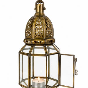 May include: A hexagonal brass lantern with clear glass panels and an ornate, cut-out dome top. The lantern features a hinged side door and a small metal ring at the top for hanging. A single lit tea light candle sits inside the base of the lantern.