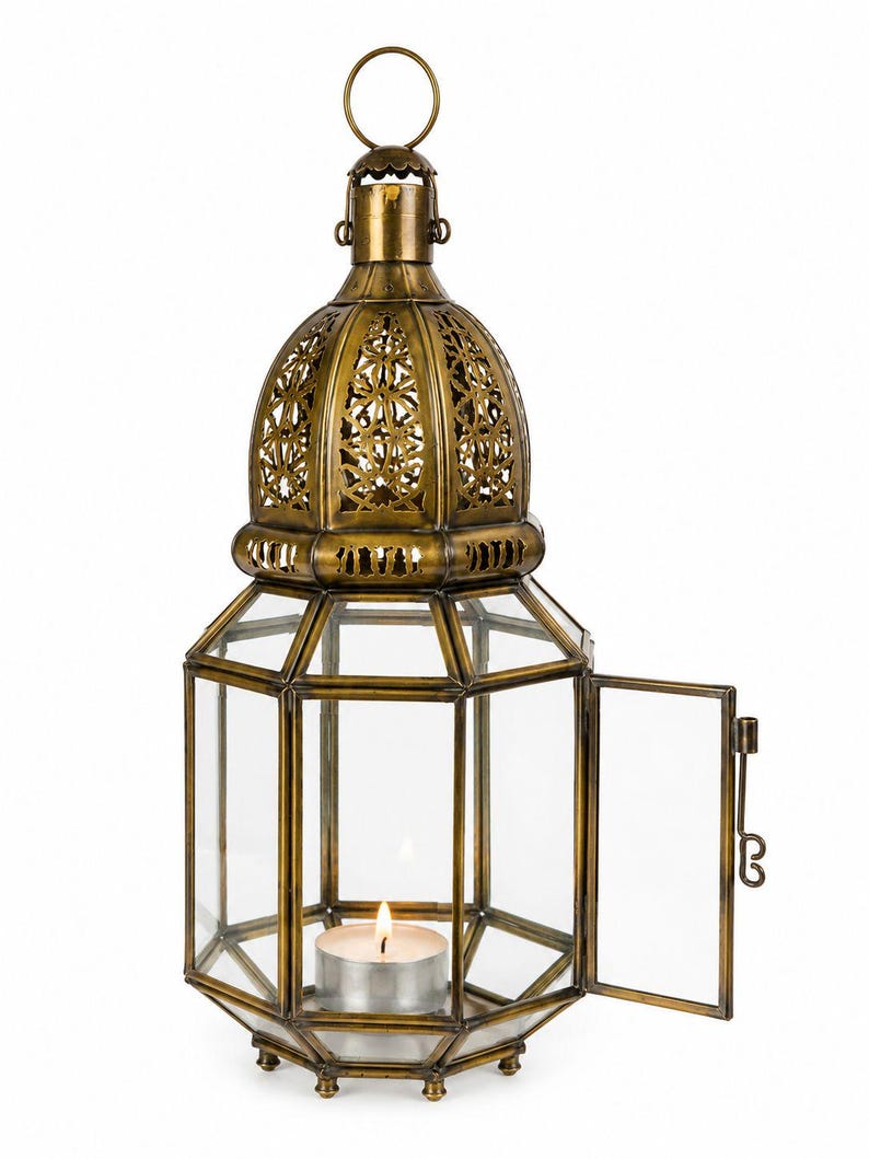 May include: A hexagonal brass lantern with clear glass panels and an ornate, cut-out dome top. The lantern features a hinged side door and a small metal ring at the top for hanging. A single lit tea light candle sits inside the base of the lantern.