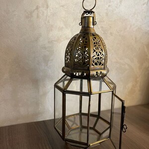 May include: A hexagonal lantern crafted from antique brass-toned metal and clear glass panels. The top features an intricate, cut-out geometric pattern and a circular hanging ring. One side panel is hinged and open, revealing the hollow interior.