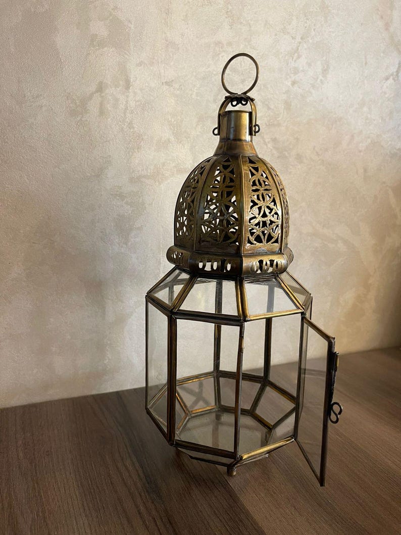 May include: A hexagonal lantern crafted from antique brass-toned metal and clear glass panels. The top features an intricate, cut-out geometric pattern and a circular hanging ring. One side panel is hinged and open, revealing the hollow interior.