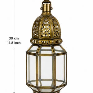 May include: A brass-colored metal lantern with clear glass panels, featuring an intricate cut-out dome top and a circular hanging ring. The lantern measures 11.8 inches in height and 5.1 inches in width, as indicated by the visible measurement markers.