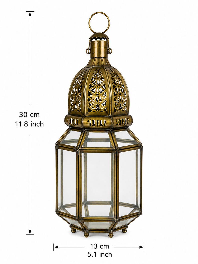 May include: A brass-colored metal lantern with clear glass panels, featuring an intricate cut-out dome top and a circular hanging ring. The lantern measures 11.8 inches in height and 5.1 inches in width, as indicated by the visible measurement markers.