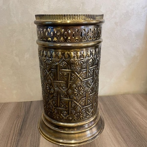 May include: A cylindrical brass container with intricate floral and geometric patterns. The container has a decorative openwork band near the top and a flared base. The overall design is ornate and detailed, with a vintage aesthetic.
