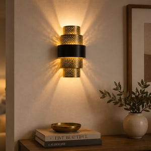 May include: A wall sconce featuring a cylindrical design with three perforated gold-toned metal sections separated by a solid black band. The light fixture emits a warm glow, casting light patterns upward and downward against the wall surface.