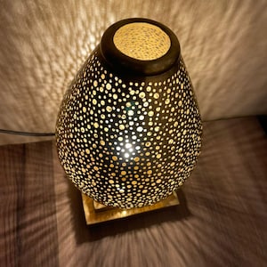May include: A decorative table lamp with a perforated metal shade. The lamp has a teardrop shape and a square base. The shade is dark brown with numerous small holes that cast a pattern of light onto the surrounding surface. The lamp is turned on, emitting a warm glow.
