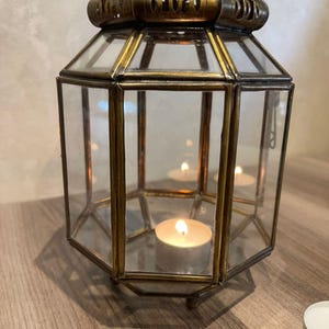 May include: A hexagonal lantern constructed with antique brass metal framing and clear glass panels. The top section features intricate cut-out metal detailing. Inside the lantern, a single lit tea light candle sits on the base, casting a warm glow.