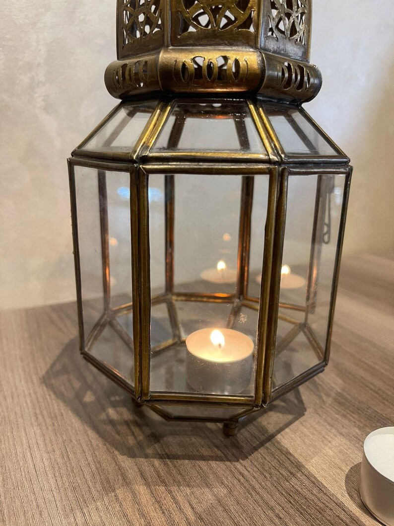 May include: A hexagonal lantern constructed with antique brass metal framing and clear glass panels. The top section features intricate cut-out metal detailing. Inside the lantern, a single lit tea light candle sits on the base, casting a warm glow.