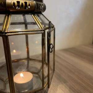 May include: A hexagonal lantern constructed with antique brass-toned metal framing and clear glass panels. The lantern features a hinged door with a simple metal latch and a decorative filigree top. A small, lit white tea light candle sits inside.