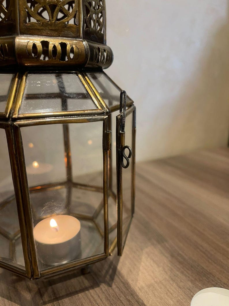 May include: A hexagonal lantern constructed with antique brass-toned metal framing and clear glass panels. The lantern features a hinged door with a simple metal latch and a decorative filigree top. A small, lit white tea light candle sits inside.