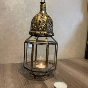 May include: A hexagonal brass lantern with clear glass panels and a decorative filigree dome top. The lantern features a small metal ring at the apex for hanging and contains a single lit white tea light candle inside, with another unlit candle nearby.