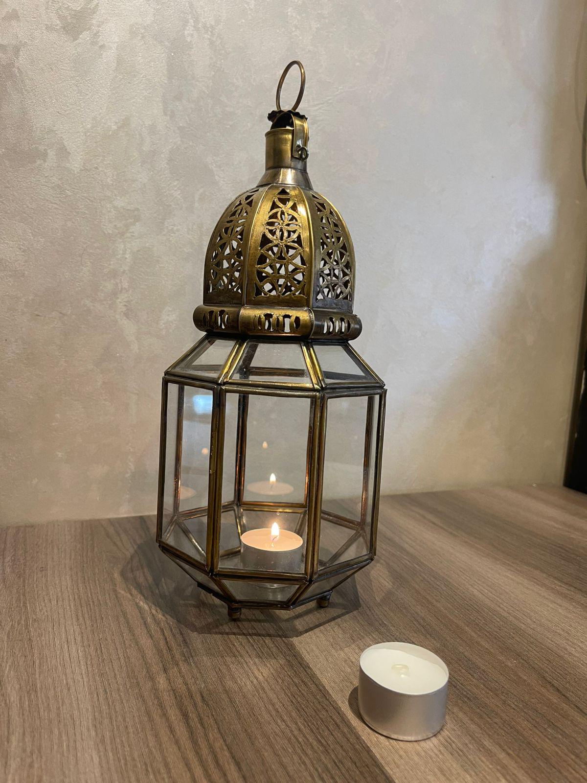 May include: A hexagonal brass lantern with clear glass panels and a decorative filigree dome top. The lantern features a small metal ring at the apex for hanging and contains a single lit white tea light candle inside, with another unlit candle nearby.