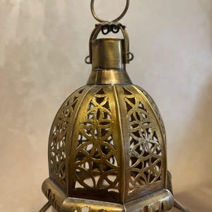 May include: A brass-colored metal lantern featuring a dome-shaped top with intricate cut-out geometric patterns. The lantern includes a circular hanging ring at the apex and a hexagonal base structure with clear glass panels framed in metal.