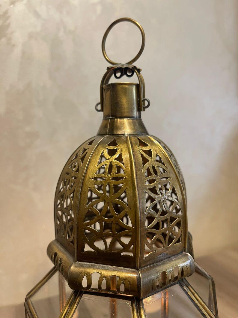 May include: A brass-colored metal lantern featuring a dome-shaped top with intricate cut-out geometric patterns. The lantern includes a circular hanging ring at the apex and a hexagonal base structure with clear glass panels framed in metal.