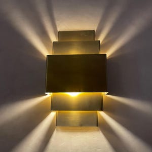 May include: A modern wall sconce with a tiered, rectangular design. The sconce is made of a brushed brass material and emits a warm, inviting light. The light casts a series of linear shadows on the wall, creating a geometric pattern. The fixture is mounted on a neutral-colored wall.
