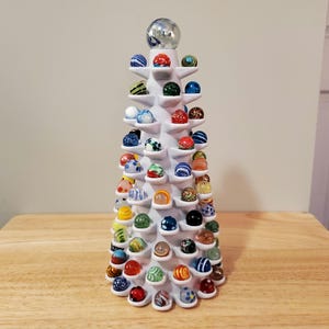 May include: A white conical display stand featuring numerous small, semi-circular shelves arranged in a spiral pattern. Each shelf holds a single colorful glass marble. The marbles vary in color, including blue, red, green, orange, and clear.