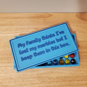 May include: A light blue marble box with a matching lid. The lid has the text "My Family thinks I've lost my marbles but I keep them in this box." The box contains several colorful marbles.