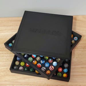 May include: Black wooden marble storage box with the word "MARBLES" embossed on the lid. The box is open, revealing a collection of colorful glass marbles in various sizes and patterns. The marbles are arranged inside the box.