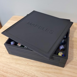 May include: A black marble storage box with the word "MARBLES" embossed on the lid. The box is partially open, revealing colorful marbles inside. The box is sitting on a wooden surface.