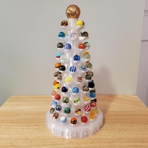 May include: A clear, tiered display stand filled with colorful glass marbles. The marbles are various sizes and feature a range of patterns and colors, including blue, red, yellow, and green. A gold-colored sphere tops the display. The stand is made of clear plastic.