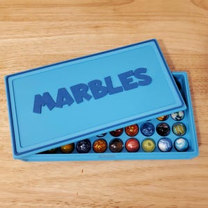 May include: A light blue marble case with the word "MARBLES" embossed on the lid. The case is open, revealing a collection of colorful marbles in various sizes and patterns. The marbles are displayed on a wooden surface.