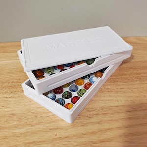 May include: A white plastic storage case with the word MARBLES embossed on the lid, containing two stacked trays filled with assorted glass marbles. The marbles feature various colors, including red, blue, green, yellow, and orange, with swirl patterns.