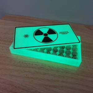 May include: A glowing green box with a hinged lid, containing marbles. The lid features a black radiation symbol and the text "RADIOACTIVE URANIUM GLASS MARBLES". The marbles are visible inside the open box.