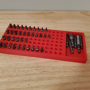 May include: A red plastic organizer with a honeycomb design holds a variety of screwdriver bits. The organizer has a separate compartment for longer bits.