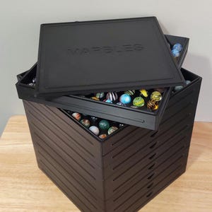 May include: A black stackable marble storage box with a lid. The lid has the word "MARBLES" embossed on it. The box is filled with colorful marbles.
