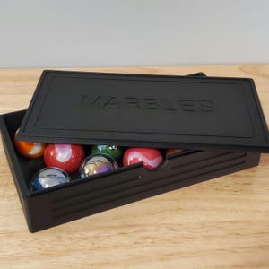 May include: A black rectangular box with a lid that says "MARBLES" on the top. The lid is open and there are colorful marbles inside.