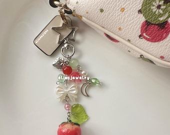 May include: A white handbag with a colorful polka dot pattern and a charm attached to the zipper pull. The charm features a star, a heart, a bow, a crescent moon, beads, and a strawberry.