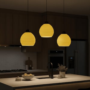 Puede incluir: Tres lámparas colgantes amarillas con un diseño texturizado en forma de panal cuelgan sobre una isla de cocina. Cada lámpara tiene una parte superior negra y un brillo cálido. La cocina cuenta con gabinetes de madera.