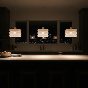 Op de afbeelding: Drie beige hanglampen met een gelaagd, cilindrisch ontwerp hangen boven een aanrecht. De lampen geven een warme gloed af. Een raam op de achtergrond onthult een stadsgezicht 's nachts.