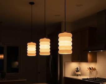 May include: Three pendant lights with textured, cylindrical shades hang above a kitchen island. The shades are a warm, off-white colour, and the cords and ceiling mounts are black. The kitchen has dark wood cabinets and a light-coloured countertop.