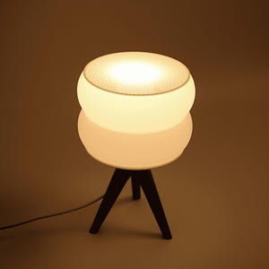 Puede incluir: Una lámpara de mesa moderna con una pantalla esférica blanca y una parte superior de malla, que descansa sobre una base circular blanca sostenida por tres patas de madera oscura. La lámpara emite un brillo cálido y suave, y se ve un cable de alimentación.
