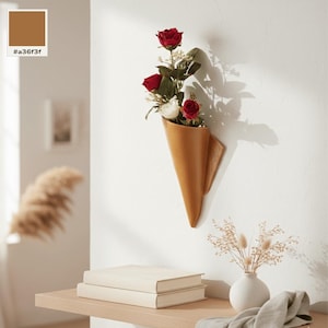 Könnte beinhalten: Eine kegelförmige Wandvase in einem warmen Braunton, die einen Strauß aus drei roten Rosen und einer weißen Rose mit grünen Blättern enthält. Die Vase ist an einer flachen Holzrückwand befestigt und an einer weißen Wand montiert.