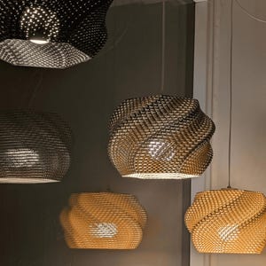 Könnte beinhalten: Mehrere Pendelleuchten mit gewebten, strukturierten Schirmen hängen von der Decke. Die Schirme sind in verschiedenen Farben gehalten, darunter Schwarz, Gold und Bronze. Die Lichter sind eingeschaltet und beleuchten das gewebte Muster.