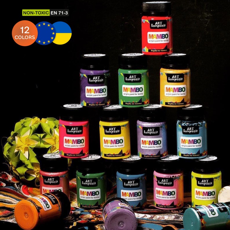Op de afbeelding: Twaalf flesjes acrylverf voor textiel in verschillende kleuren, met het label "ART kompozit MAMBO". De flesjes zijn in een piramidevorm op een zwarte achtergrond gerangschikt. Het label vermeldt ook "NON-TOXIC EN 71-3" en "12 KLEUREN".