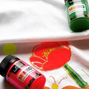 May include: Two bottles of fabric paint, one green and one pink, with the brand name "MAMBO" and the text "acrylic paint for textile" on the labels. The bottles are on a white fabric with painted flowers.