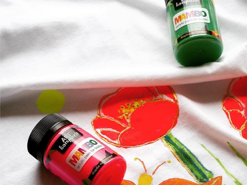 May include: Two bottles of fabric paint, one green and one pink, with the brand name "MAMBO" and the text "acrylic paint for textile" on the labels. The bottles are on a white fabric with painted flowers.