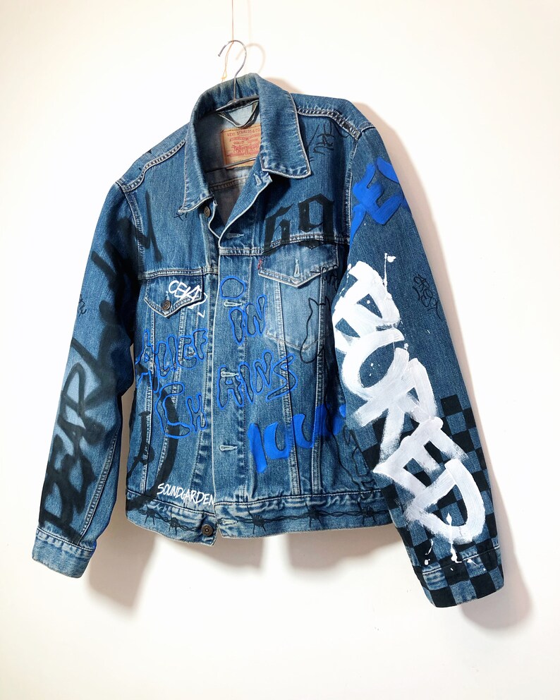 May include: A blue denim jacket with black and white graffiti and blue and white lettering. The lettering includes the words "SOUNDGARDEN", "HOME", "FOR", "ALWAYS", "NOW", and "WORKED".